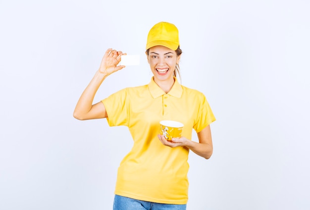 Chica femenina en uniforme amarillo entregando una taza de fideos para llevar amarilla y presentando su tarjeta de visita al cliente.