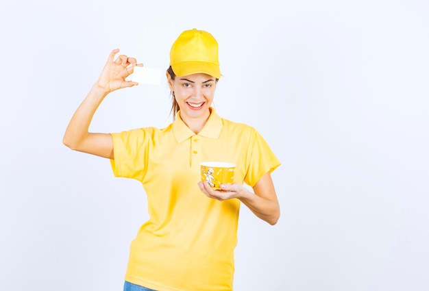 Chica femenina en uniforme amarillo entregando una taza de fideos para llevar amarilla y presentando su tarjeta de visita al cliente.
