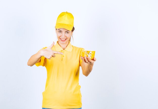 Chica femenina en uniforme amarillo entregando una taza de fideos para llevar amarillo al cliente.