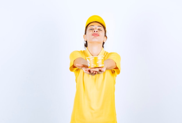 Chica femenina en uniforme amarillo entregando una taza de fideos para llevar amarillo al cliente.