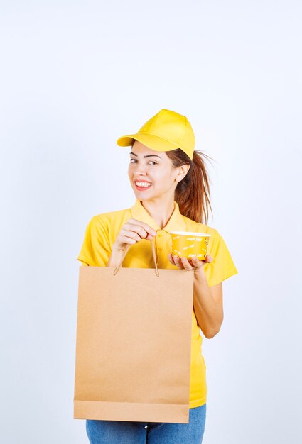 Chica femenina en uniforme amarillo sosteniendo una bolsa de compras y una taza amarilla de fideos para llevar.