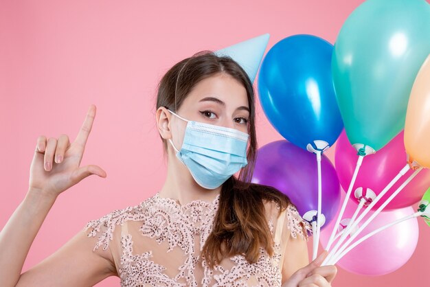 Chica de fiesta de vista frontal de primer plano con corona y máscara sosteniendo globos haciendo pistola de dedo