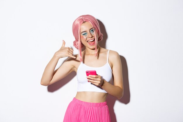 Chica fiestera alegre haciendo señal de llamada telefónica y guiñando un ojo coqueto a la cámara, sosteniendo el teléfono inteligente, de pie con peluca rosa.