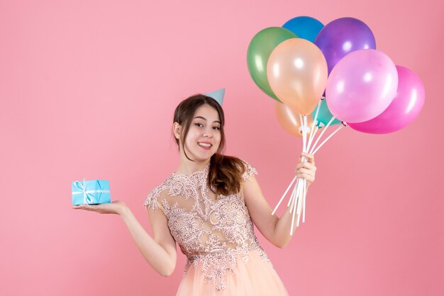 Chica fiestera feliz con gorro de fiesta sosteniendo globos de colores y regalo en rosa