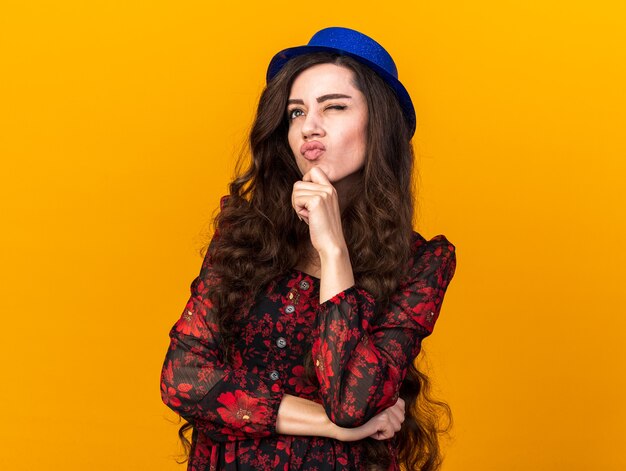 Chica fiestera joven pensativa con sombrero de fiesta mirando hacia arriba manteniendo la mano en la barbilla frunciendo los labios con un ojo cerrado aislado en la pared naranja con espacio de copia
