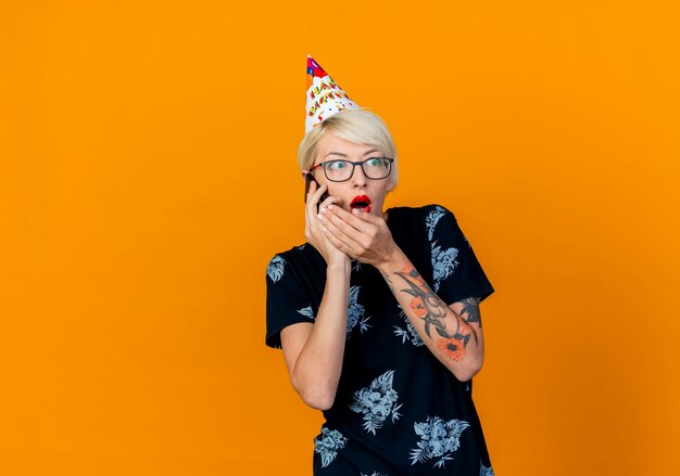 Chica fiestera rubia joven sorprendida con gafas y gorra de cumpleaños hablando por teléfono manteniendo la mano cerca de la boca mirando al lado aislado sobre fondo naranja con espacio de copia