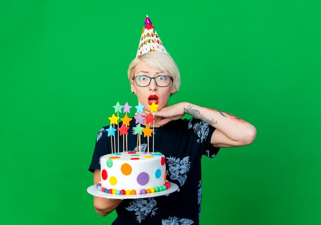 Chica fiestera rubia joven sorprendida con gafas y gorro de cumpleaños sosteniendo pastel de cumpleaños con estrellas manteniendo la mano debajo de la barbilla mirando a cámara aislada sobre fondo verde con espacio de copia