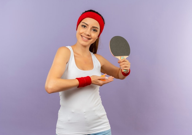 Chica fitness joven alegre en ropa deportiva con diadema sosteniendo raqueta y pelota de tenis de mesa con una sonrisa en la cara de pie sobre la pared gris