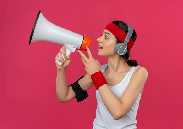 Chica fitness joven disgustado en ropa deportiva con diadema y auriculares gritando al megáfono parado sobre pared rosa