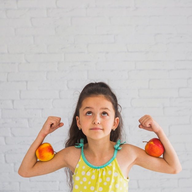 Foto gratuita chica flexionando sus manos sosteniendo la manzana en su bíceps mirando hacia arriba