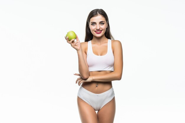 Chica en forma y deportiva en ropa interior blanca. Mujer hermosa y sana que come la manzana verde sobre la pared gris.