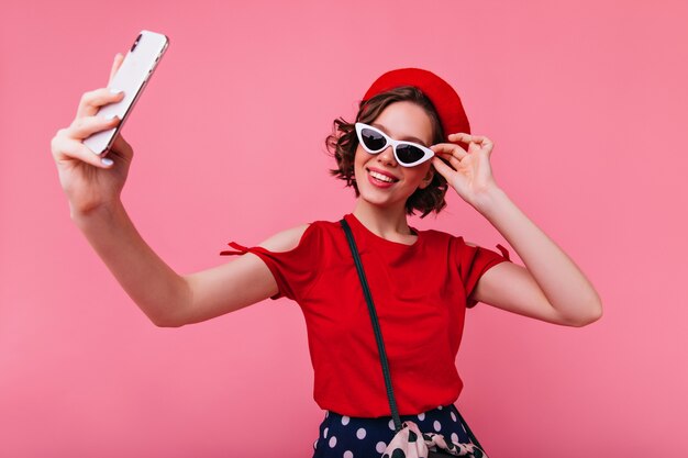 Chica francesa elegante con tatuajes haciendo selfie. Elegante mujer blanca con boina y gafas de sol tomando una foto de sí misma.