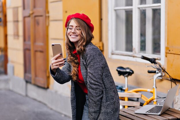 Chica francesa rizada refinada haciendo selfie cerca de la mesa de madera con un portátil en él