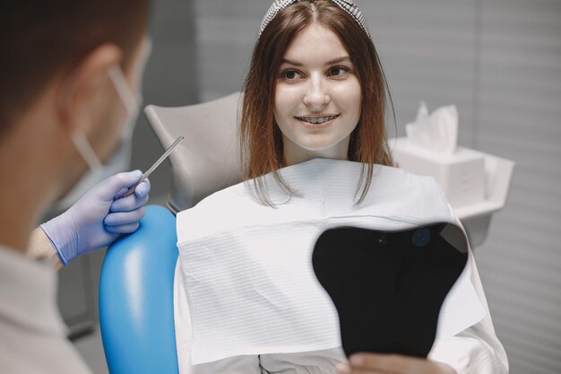 Chica con frenillos mirando en el espejo a sí misma en el consultorio del dentista. Estomatólogo con guantes azules y mascarilla