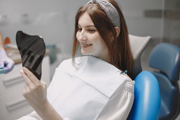 Chica con frenillos mirando en el espejo a sí misma en el consultorio del dentista. La silla es azul