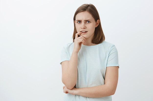 Chica frente a decisiones difíciles pensando y dudando mordiendo el dedo mientras fruncía el ceño luciendo intenso y perplejo teniendo problemas y sin un plan en mente, posando preocupada contra la pared gris