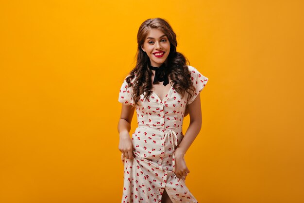 Chica fresca en vestido estampado cereza posando sobre fondo naranja. Mujer de moda ondulada en ropa blanca sonriendo sobre fondo aislado.