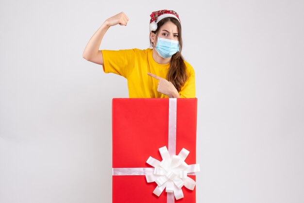 Chica fuerte con gorro de Papá Noel mostrando músculo de pie detrás de un gran regalo de Navidad en blanco