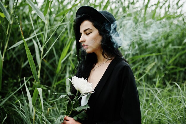 Chica fumadora sensual toda en labios rojos negros y sombrero Mujer dramática gótica sostiene flor de crisantemo blanco y fuma en caña común
