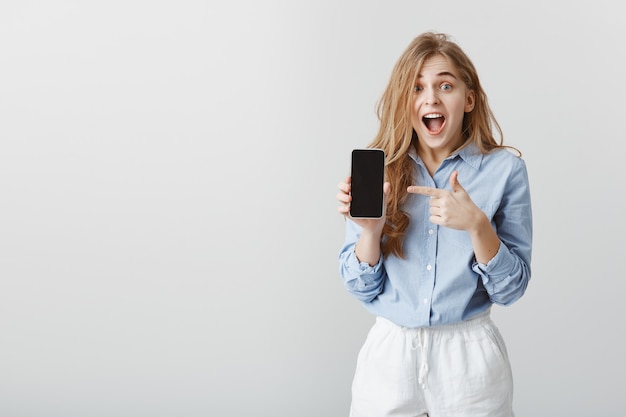 Chica ganó smartphone en lotería. Retrato de mujer joven encantadora asombrada en blusa azul mostrando teléfono inteligente y apuntando al dispositivo con el dedo índice, dejando caer la mandíbula, gritando de emoción y sorpresa