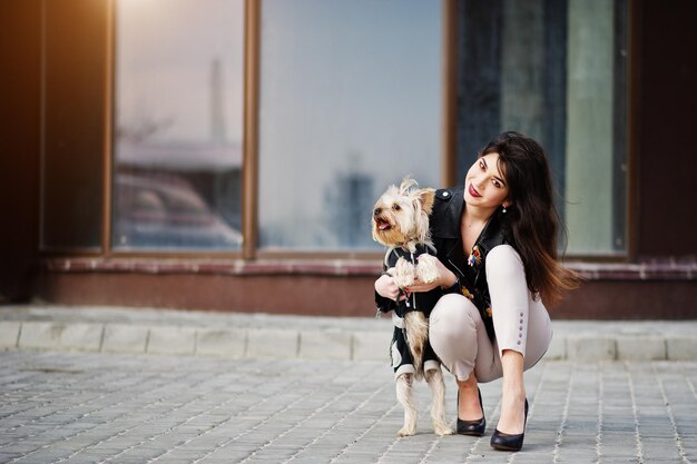 Chica gitana morena con perro yorkshire terrier posó contra grandes ventanales Casa modelo usa chaqueta de cuero y camiseta con pantalones de adorno y zapatos con tacones altos