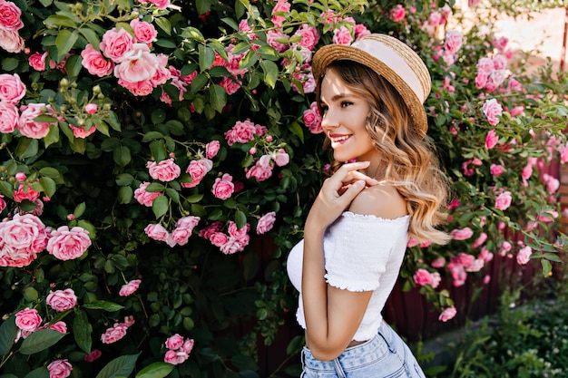 Chica glamorosa en camiseta blanca posando en el hermoso jardín. Disparo al aire libre de dama de moda con sombrero disfrutando de sabor a rosas.