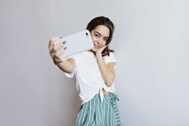 Chica glamorosa de ojos oscuros con smartphone para selfie. adorable mujer morena tomando una foto de sí misma.