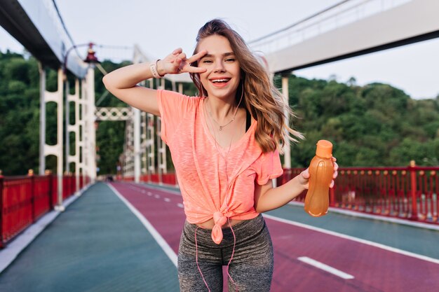 Chica glamorosa posando después de maratón con jugo. Riendo a una mujer bonita de pie en la pista de ceniza con el signo de la paz.