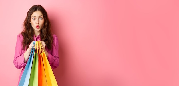 Foto gratuita chica glamurosa sosteniendo bolsas de compras con los labios fruncidos y luciendo tímida de pie contra el fondo rosa