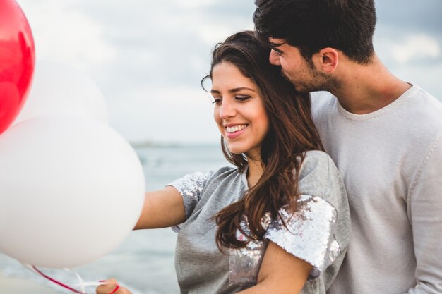 Chica con globos mientras su novio la abraza con el mar de fondo