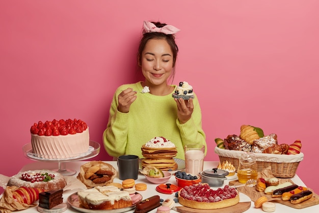 Chica golosa hambrienta sostiene cupcake en una mano, cuchara con crema en la otra, obtiene porción de azúcar disfruta de un delicioso refrigerio durante el evento festivo mantiene los ojos cerrados por placer.