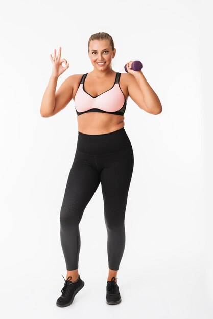 Chica gorda sonriente con top deportivo y polainas sosteniendo pesas en la mano felizmente mostrando un gesto correcto mientras mira a la cámara sobre fondo blanco