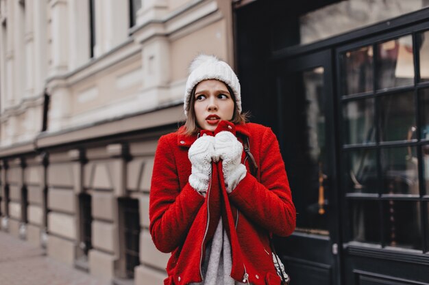 Chica con gorro de punto blanco como la nieve y guantes está temblando de frío, envolviéndose en un abrigo rojo.