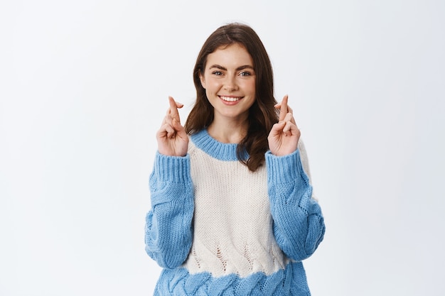 Chica con grandes esperanzas pidiendo deseos, cruzando los dedos, buena suerte y sonriendo, rezando o esperando un resultado positivo, de pie contra la pared blanca