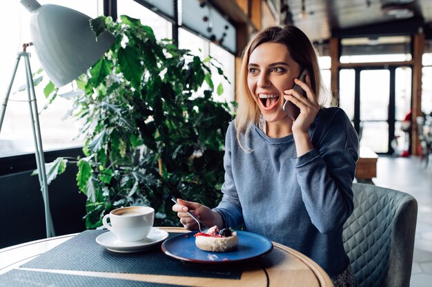 chica gratamente sorprendida teniendo conversación telefónica, comiendo postre y tomando café