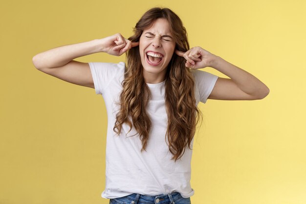 Chica gritando molesta demanda dejar de tocar la guitarra. Mujer irritada molesta cerrar los ojos gritando tapón decepcionado dedos índices agujeros para los oídos disgustado fuerte ruido terrible atril fondo amarillo