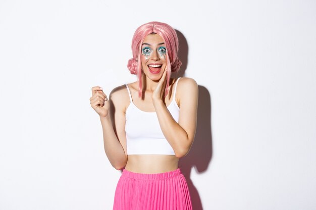 Chica guapa alegre con maquillaje brillante, vestida para celebración navideña o fiesta, mostrando tarjeta de crédito y sonriendo emocionada, de pie.