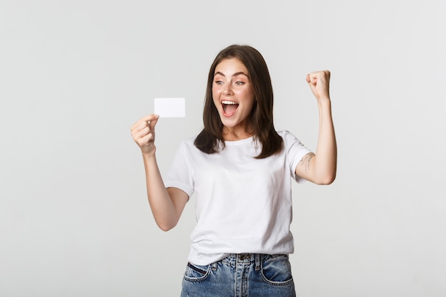 Chica guapa alegre regocijándose y mirando la tarjeta de crédito, bomba de puño mientras triunfa, blanco.