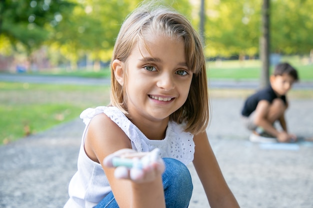 Chica guapa alegre sentada y dibujando sobre hormigón, sosteniendo coloridos trozos de tizas en la mano. Fotografía de cerca. Concepto de infancia y creatividad.