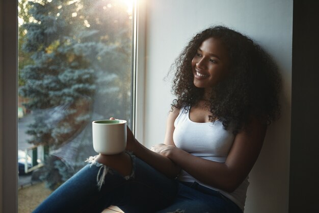 Chica guapa de apariencia de raza mixta sosteniendo una taza grande y mirando a través de la ventana con una sonrisa alegre, mirando algo agradable afuera, tomando té o café. Personas y estilo de vida
