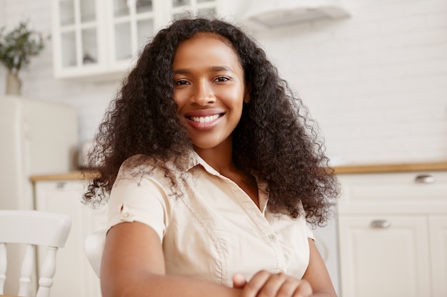 Chica guapa atractiva de origen afroamericano que expresa buenas emociones positivas, sentada contra el elegante interior de la cocina, con una sonrisa radiante y dentuda. Concepto de etnia y belleza