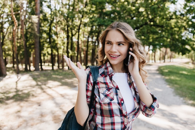 Chica guapa de buen humor llamando amigo mientras camina en el parque. Tiro al aire libre de mujer rizada agradable posando con mochila y teléfono.