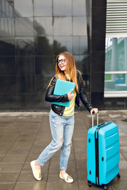 Chica guapa con cabello largo está caminando con maleta afuera en el aeropuerto. Viste chaqueta negra con jeans y sostiene una computadora portátil. Ella luce feliz.