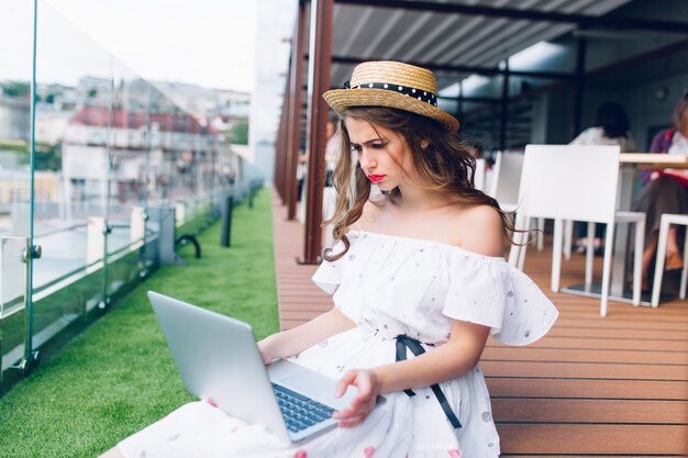 Chica guapa con cabello largo está sentada en el piso de la terraza. Lleva un vestido blanco con hombros desnudos, lápiz labial rojo y sombrero. Ella tiene una computadora portátil en las rodillas y parece molesta.