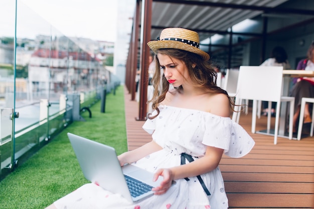 Chica guapa con cabello largo está sentada en el piso de la terraza. Lleva un vestido blanco con hombros desnudos, lápiz labial rojo y sombrero. Ella tiene una computadora portátil en las rodillas y parece molesta.