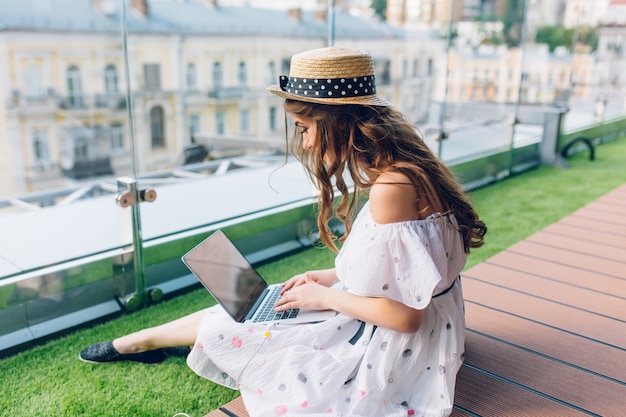 Chica guapa con cabello largo está sentada en el piso de la terraza. Lleva un vestido blanco con hombros desnudos y sombrero. Ella está escribiendo en la computadora portátil de rodillas.