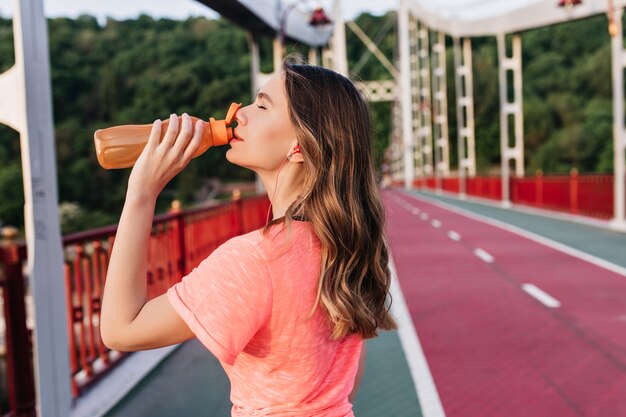 Chica guapa con cabello ondulado bebiendo agua después de maratón. Señora caucásica refinada posando en la pista de ceniza durante el entrenamiento.