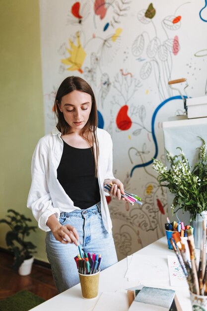 Chica guapa cerca del escritorio sosteniendo soñadoramente rotuladores de colores en la mano con grandes patrones de lienzo en el fondo en casa