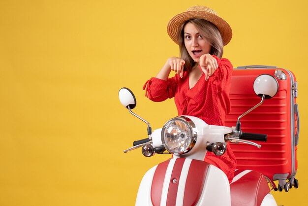 Chica guapa en ciclomotor con maleta roja apuntando a la cámara