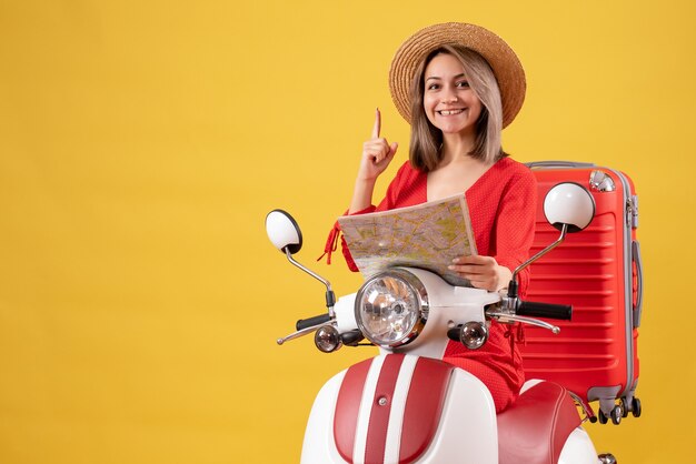 Chica guapa en ciclomotor con maleta roja sosteniendo el mapa apuntando con el dedo hacia arriba